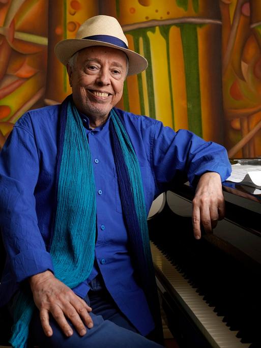 Sergio Mendes sitzt mit Hut an einem Flügel, im Hintergrund ein buntes Gemälde. Er lacht in die Kamer. 