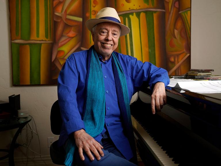 Sergio Mendes sitzt mit Hut an einem Flügel, im Hintergrund ein buntes Gemälde. Er lacht in die Kamer. 