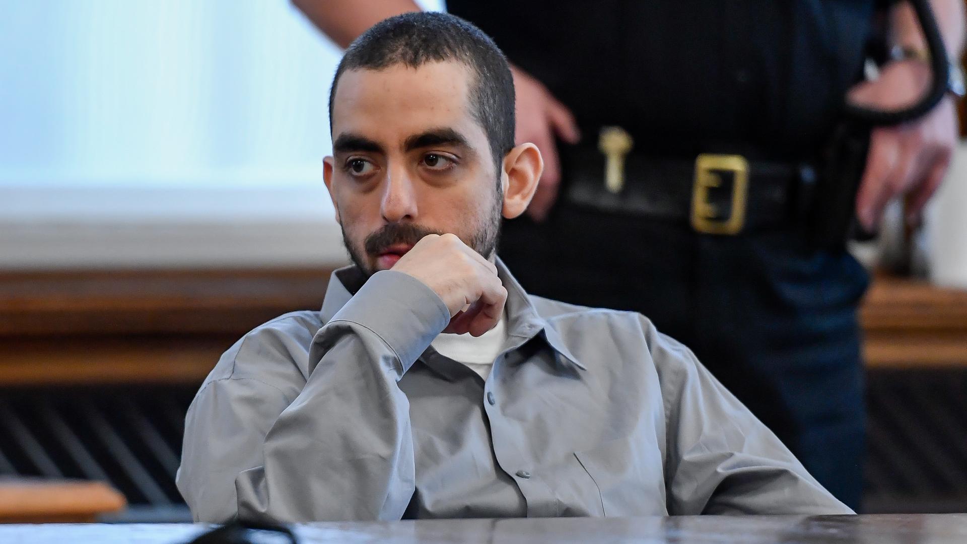Der Attentäter Hadi Matar sitzt in grauer Häftlingskleidung an einem Tisch und hält die rechte Hand ans Kinn. Er hat einen Vollbart und schaut zur Seite. Dahinter unscharf im Anschnitt ein Polizist. Der Attentäter Hadi Matar sitzt in grauer Häftlingskleidung an einem Tisch und hält die rechte Hand ans Kinn. Er hat einen Vollbart und schaut zur Seite. Dahinter unscharf im Anschnitt ein Polizist.