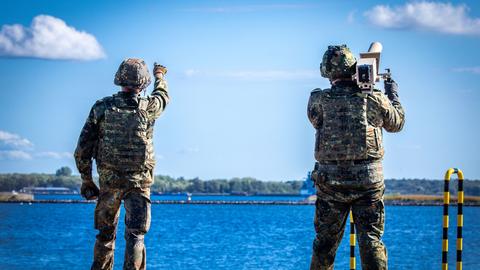 Zwei Soldaten mit Helm und Tarnkleidung stehen an einem Hafen und blicken in den Himmel, einer von ihnen trägt einen Störsender auf der Schulter. 
