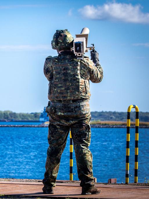 Zwei Soldaten mit Helm und Tarnkleidung stehen an einem Hafen und blicken in den Himmel, einer von ihnen trägt einen Störsender auf der Schulter. Zwei Soldaten mit Helm und Tarnkleidung stehen an einem Hafen und blicken in den Himmel, einer von ihnen trägt einen Störsender auf der Schulter.