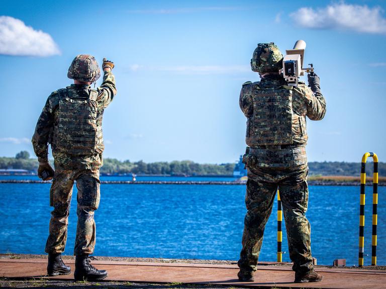 Zwei Soldaten mit Helm und Tarnkleidung stehen an einem Hafen und blicken in den Himmel, einer von ihnen trägt einen Störsender auf der Schulter. Zwei Soldaten mit Helm und Tarnkleidung stehen an einem Hafen und blicken in den Himmel, einer von ihnen trägt einen Störsender auf der Schulter.
