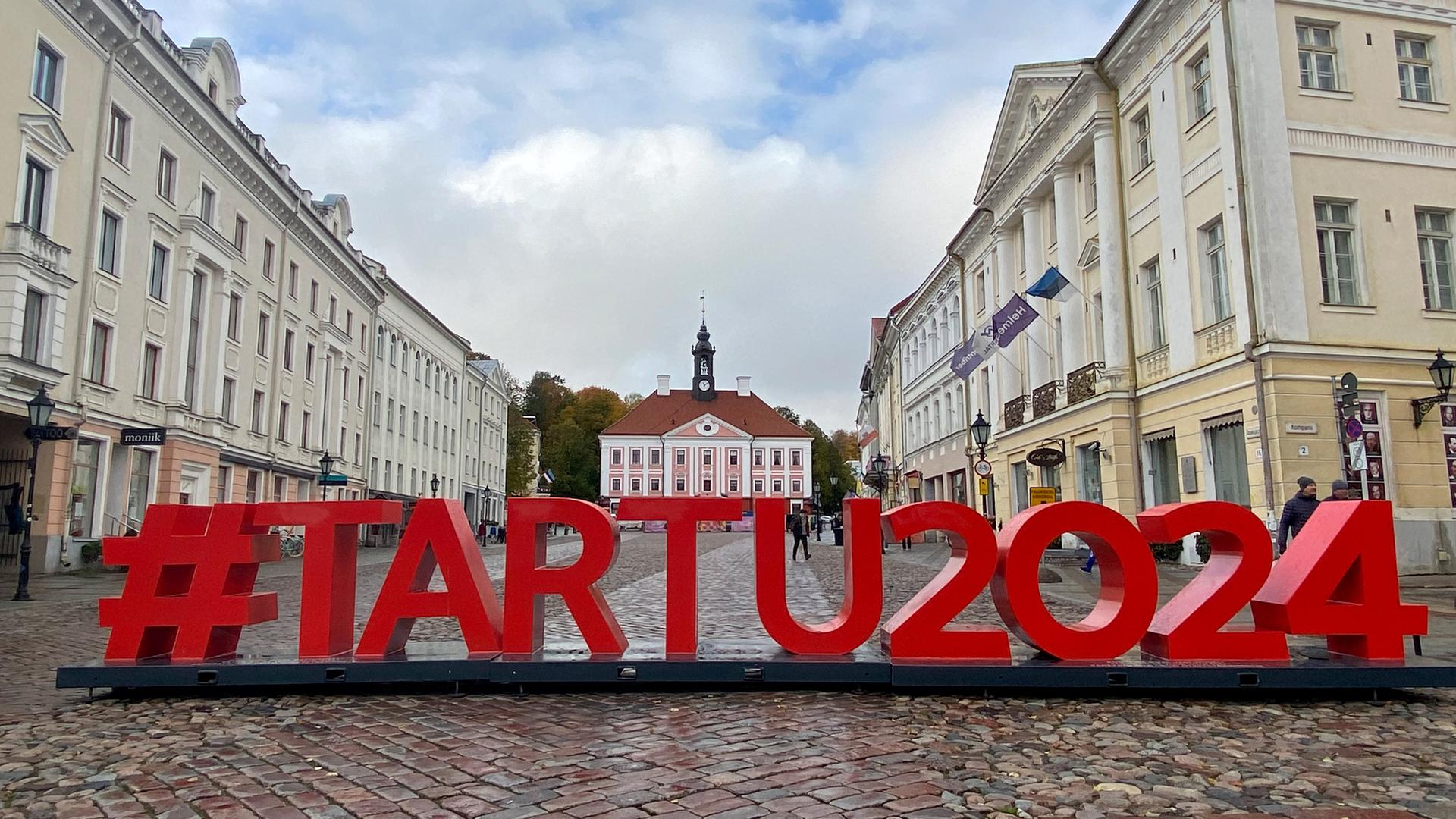 Ein Schriftzug "#Tartu2024" ist in Großbuchstaben auf dem gepflasterten Rathausplatz von Tartu aufgestellt. Im Hintergrund sieht man das historische Rathaus