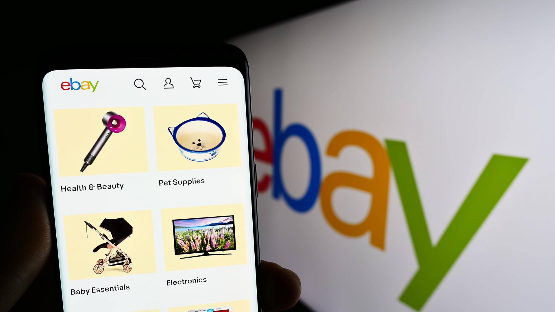 Eine Person hält ein Smartphone mit der Ebay-App. Im Hintergrund das Ebay-Logo.