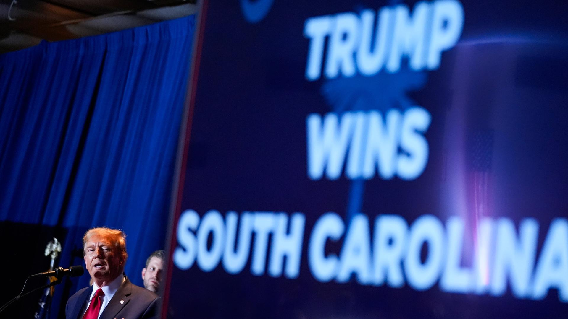 Donald Trump steht triumphierend auf einer Bühne vor Anhängern an einem Redepult. Im Hintergrund ist ein riesiger Schriftzug an eine Wand projiziert, der seinen Sieg in der Vorwahl der Republikaner im US-Bundesstaat South-Carolina verkünde.