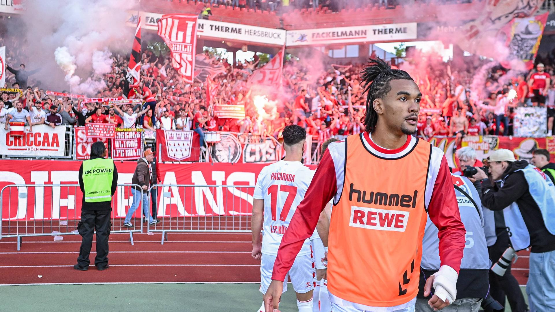 Ein Spieler des 1. FC Köln im Stadion. Hinter ihm sind auf den Rängen Fans, die Pyrotechnik abbrennen. 