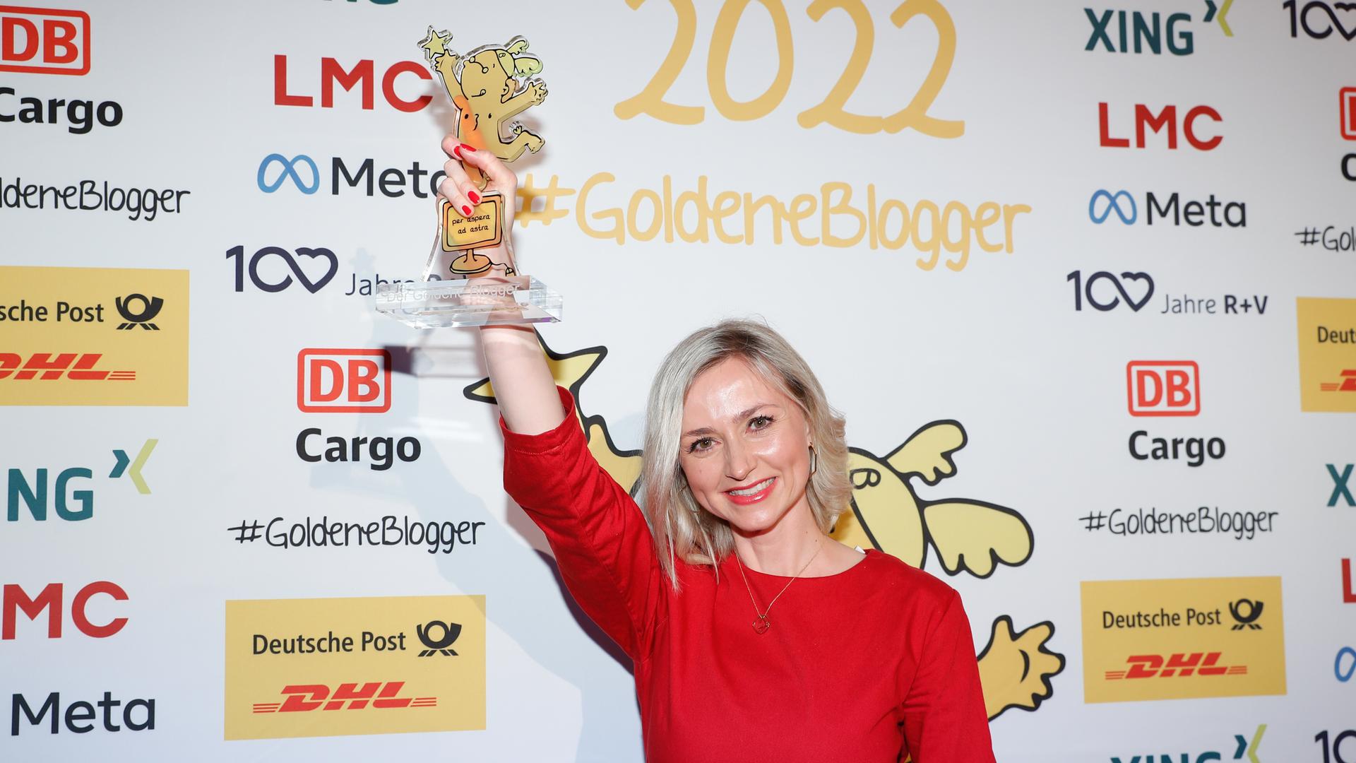 Eine blonde Frau in einem roten Kleid reckt eine Trophäe in die Höhe. Auf der Wand hinter ihr steht "Goldene Blogger". Eine blonde Frau in einem roten Kleid reckt eine Trophäe in die Höhe. Auf der Wand hinter ihr steht "Goldene Blogger".