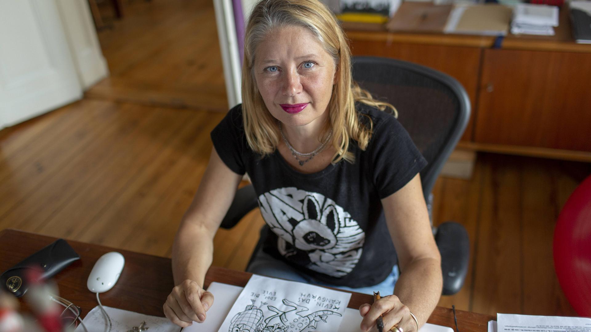 Kat Menschik sitzt an ihrem Schreibtisch. Sie hat blondes, schulterlanges Haar und trägt roten Lippenstift. Vor ihr liegt eine Zeichung in Schwarzweiß. Kat Menschik sitzt an ihrem Schreibtisch. Sie hat blondes, schulterlanges Haar und trägt roten Lippenstift. Vor ihr liegt eine Zeichung in Schwarzweiß.