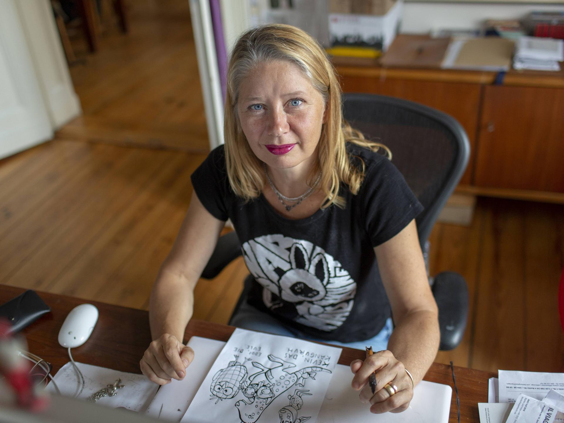 Kat Menschik sitzt an ihrem Schreibtisch. Sie hat blondes, schulterlanges Haar und trägt roten Lippenstift. Vor ihr liegt eine Zeichung in Schwarzweiß.