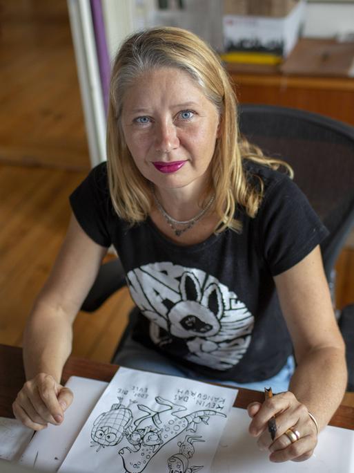 Kat Menschik sitzt an ihrem Schreibtisch. Sie hat blondes, schulterlanges Haar und trägt roten Lippenstift. Vor ihr liegt eine Zeichung in Schwarzweiß.