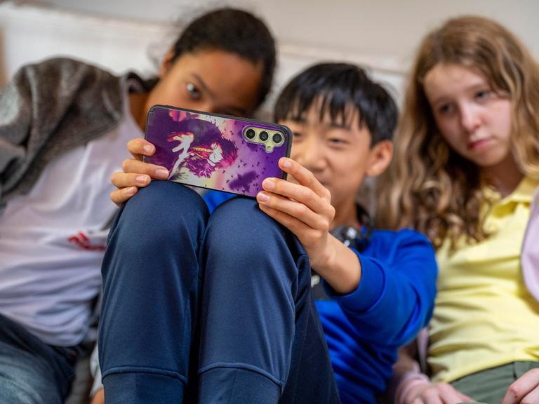 Zwei Mädchen und ein Junge sitzen dicht nebeneinander auf der Couch und machen mit einem Smartphone ein Selfie von sich