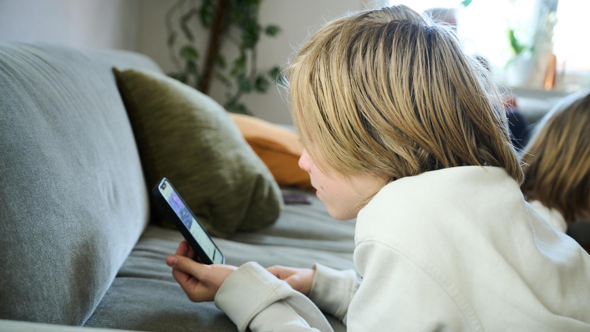 Ein Junge liegt auf einem Sofa und guckt auf sein Smartphone. (gestellte Szene)