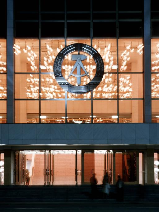 Blick von außen in den hell erleuchteten Palast der Republik bei Nacht. An der Glasfassade hängt das Staatswappen der DDR: ein Ährenkranz, in dem sich ein Hammer und ein Zirkel befinden