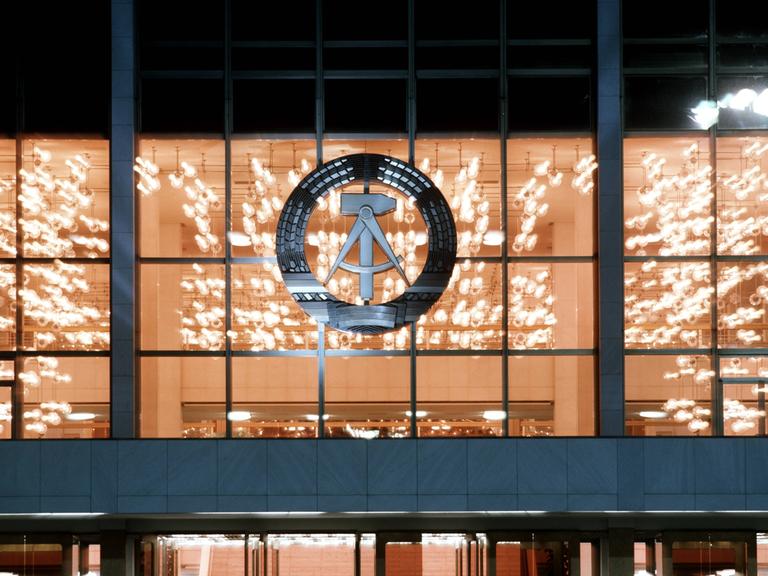 Blick von außen in den hell erleuchteten Palast der Republik bei Nacht. An der Glasfassade hängt das Staatswappen der DDR: ein Ährenkranz, in dem sich ein Hammer und ein Zirkel befinden