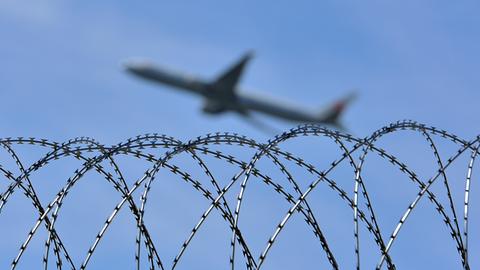 Über einem Natozaun fliegt, in der Unschärfe, ein Flugzeug vor blauem Himmel.