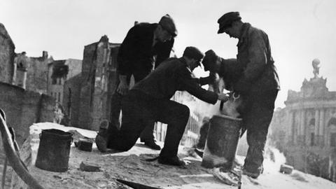 Das Schwarz-Weiß-Foto zeigt drei Männer vor beschädigten Fassaden im Jahr 1947 in der Berliner Mauerstraße. Sie hantieren mit Metalleimern und bereiten die Sprengung des Bunkers der Gauleitung der NSDAP vor. Er war in die Ruine der Dreifaltigkeitskirche eingebaut worden.