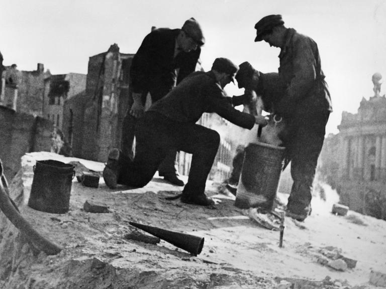 Das Schwarz-Weiß-Foto zeigt drei Männer vor beschädigten Fassaden im Jahr 1947 in der Berliner Mauerstraße. Sie hantieren mit Metalleimern und bereiten die Sprengung des Bunkers der Gauleitung der NSDAP vor. Er war in die Ruine der Dreifaltigkeitskirche eingebaut worden.