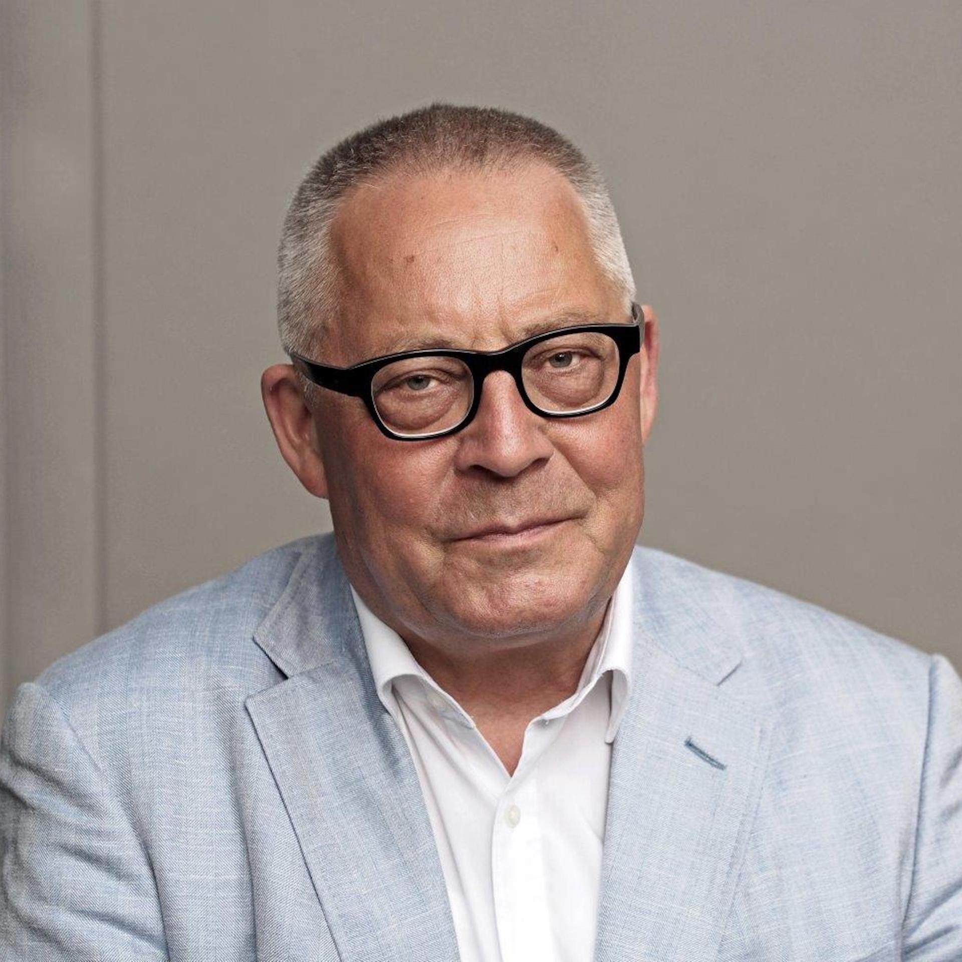 Ein Porträt von Karl Schlögel. Er sitzt in weißem Hemd und hellblauem Sakko vor einer grauen Wand und schaut in die Kamera.