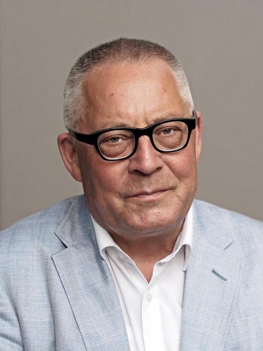 Ein Porträt von Karl Schlögel. Er sitzt in weißem Hemd und hellblauem Sakko vor einer grauen Wand und schaut in die Kamera.