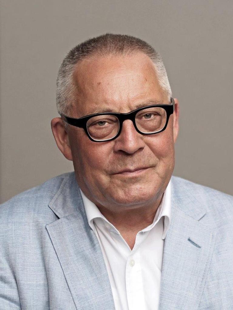 Ein Porträt von Karl Schlögel. Er sitzt in weißem Hemd und hellblauem Sakko vor einer grauen Wand und schaut in die Kamera.