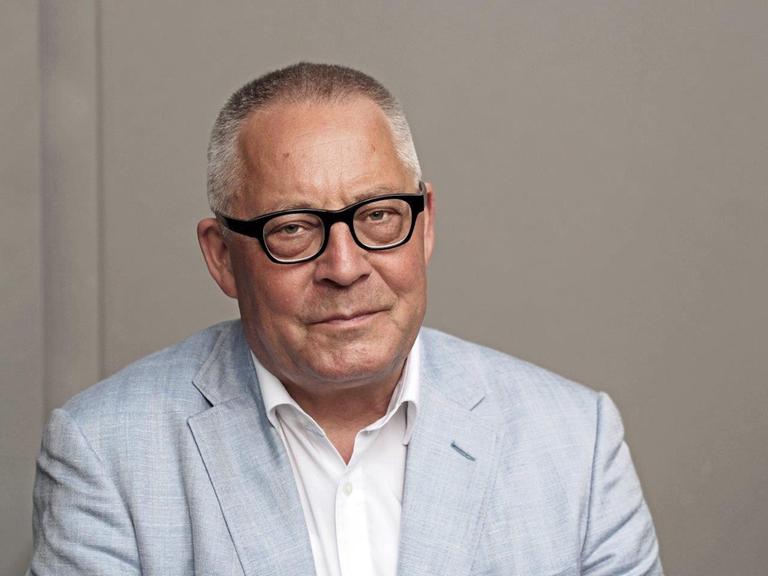 Ein Porträt von Karl Schlögel. Er sitzt in weißem Hemd und hellblauem Sakko vor einer grauen Wand und schaut in die Kamera.