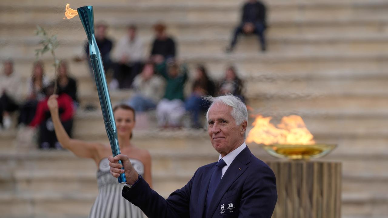 Malago hält die Fackel in die Höhe; hinter ihm eine Frau in einem römisch anmutenden Kostüm, die einen Plivenzweig in die Höhe hält. Im Hintergrund eine größere Flamme sowie steinerne Sitzreihen, auf denen ein paar Zuschauer sitzen.