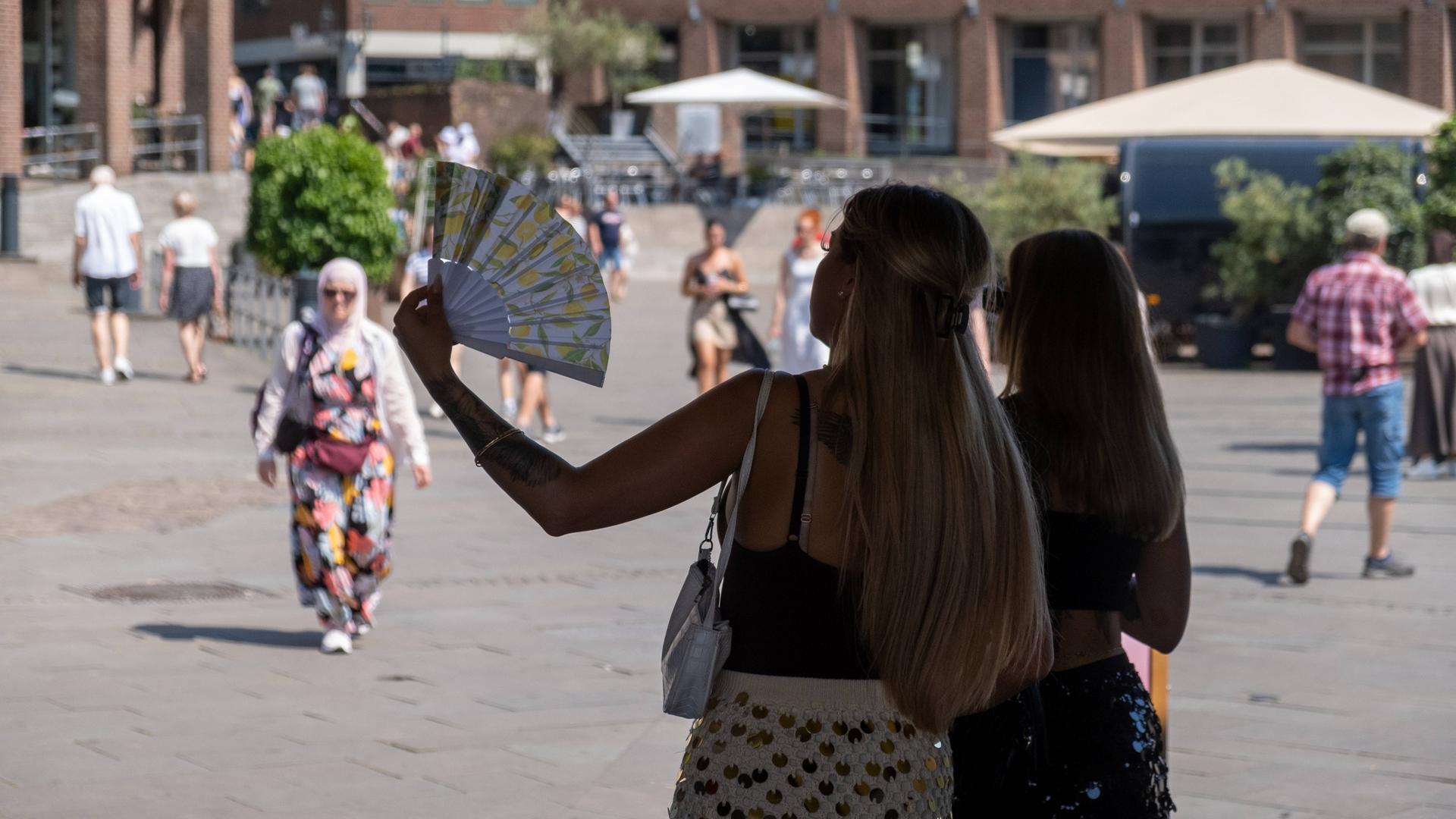 Zwei Personen stehen im Schatten vor einem Platz auf dem Menschen umhergehen. Eine der Personen hält einen Fächer in der Hand und fächelt Luft.
