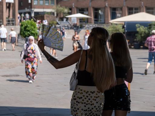 Zwei Personen stehen im Schatten vor einem Platz auf dem Menschen umhergehen. Eine der Personen hält einen Fächer in der Hand und fächelt Luft.