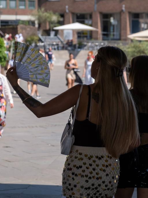 Zwei Personen stehen im Schatten vor einem Platz auf dem Menschen umhergehen. Eine der Personen hält einen Fächer in der Hand und fächelt Luft.