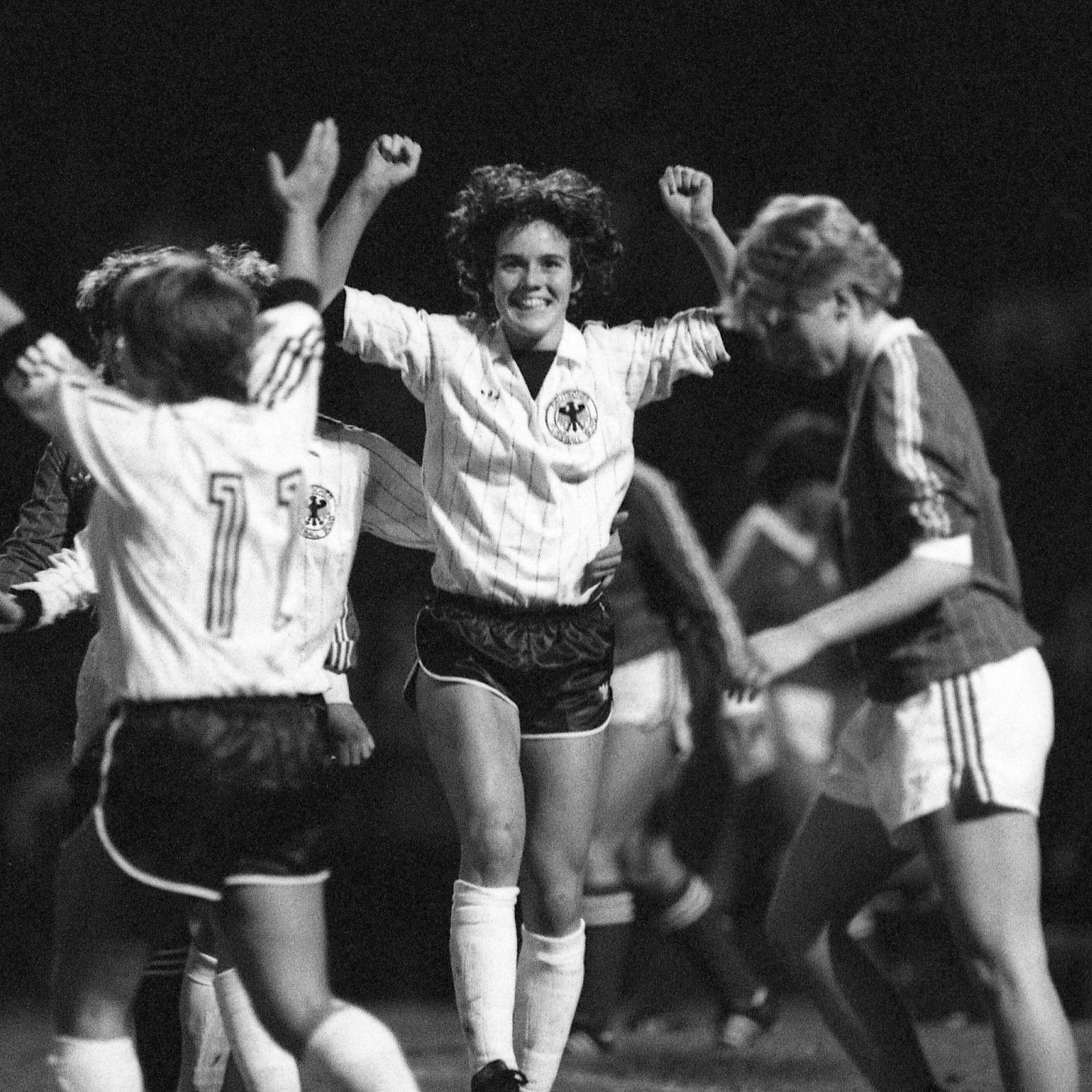 Ein schwarz-weiß-Foto zeigt mehrere Fußballspielerinnen auf dem Feld. Eine Spielerin hebt jubelnd die Arme. Die Szene stammt aus einem Frauen-Länderspiel 1982.