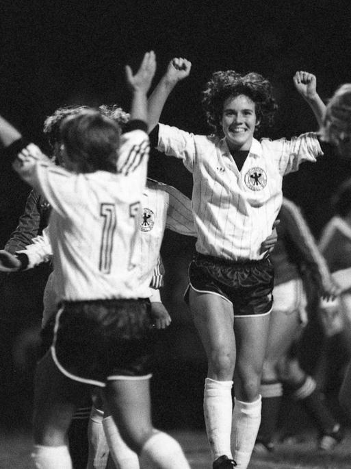 Ein schwarz-weiß-Foto zeigt mehrere Fußballspielerinnen auf dem Feld. Eine Spielerin hebt jubelnd die Arme. Die Szene stammt aus einem Frauen-Länderspiel 1982.