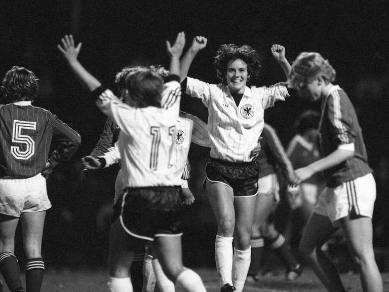 Ein schwarz-weiß-Foto zeigt mehrere Fußballspielerinnen auf dem Feld. Eine Spielerin hebt jubelnd die Arme. Die Szene stammt aus einem Frauen-Länderspiel 1982.