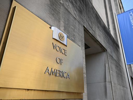 Blick von schräg unten auf das Schild am Eingang des Gebäudes von Voice of America, im hintergrund weht eine bleue Fahne mit dem Logo