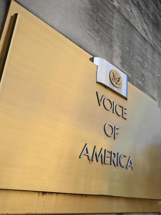 Blick von schräg unten auf das Schild am Eingang des Gebäudes von Voice of America, im hintergrund weht eine bleue Fahne mit dem Logo