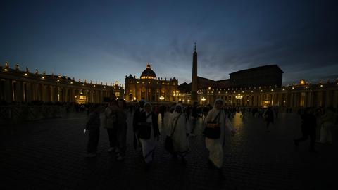Gläubige verlassen den Petersplatz; die Kuppel des Petersdoms wird beleuchtet.