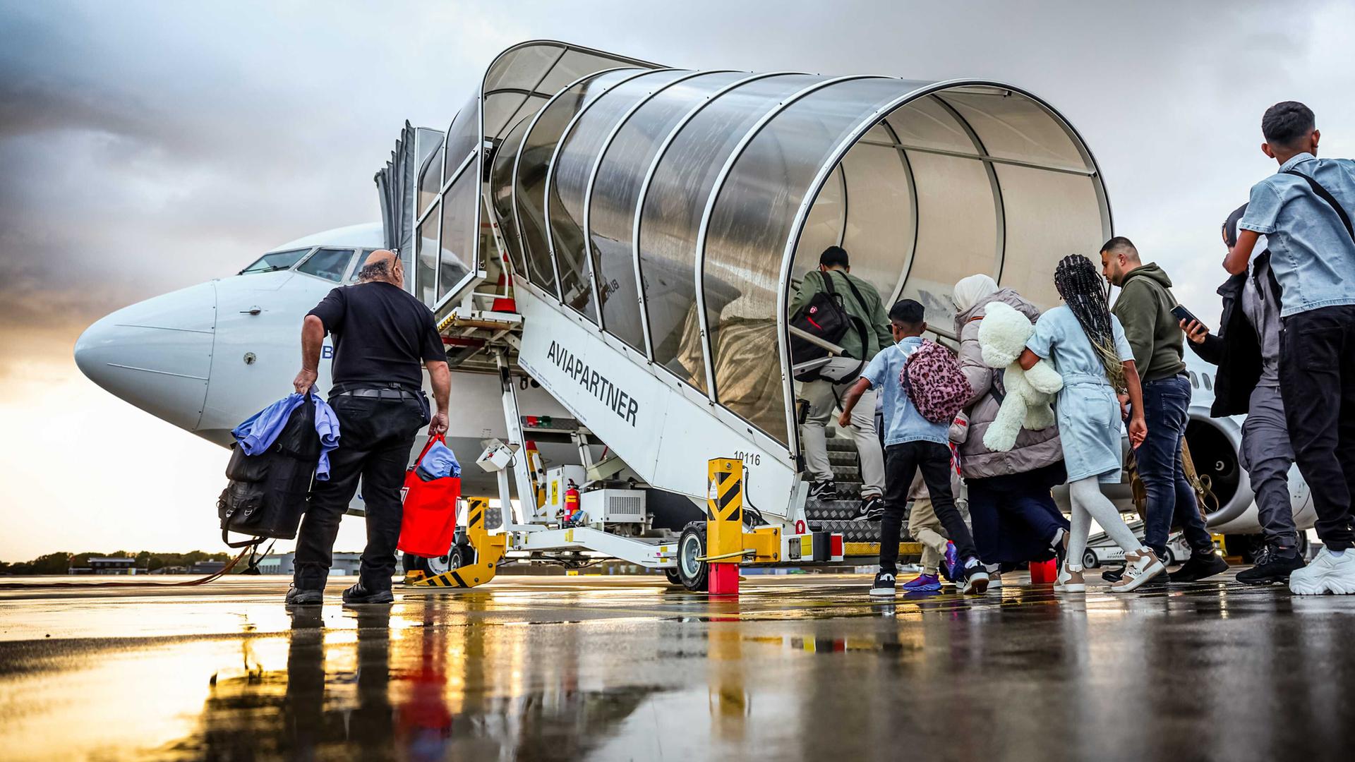 Blick auf eine Schlange von Menschen, die auf einem Rollfeld über eine überdachte Treppe in ein Flugzeug steigen. 