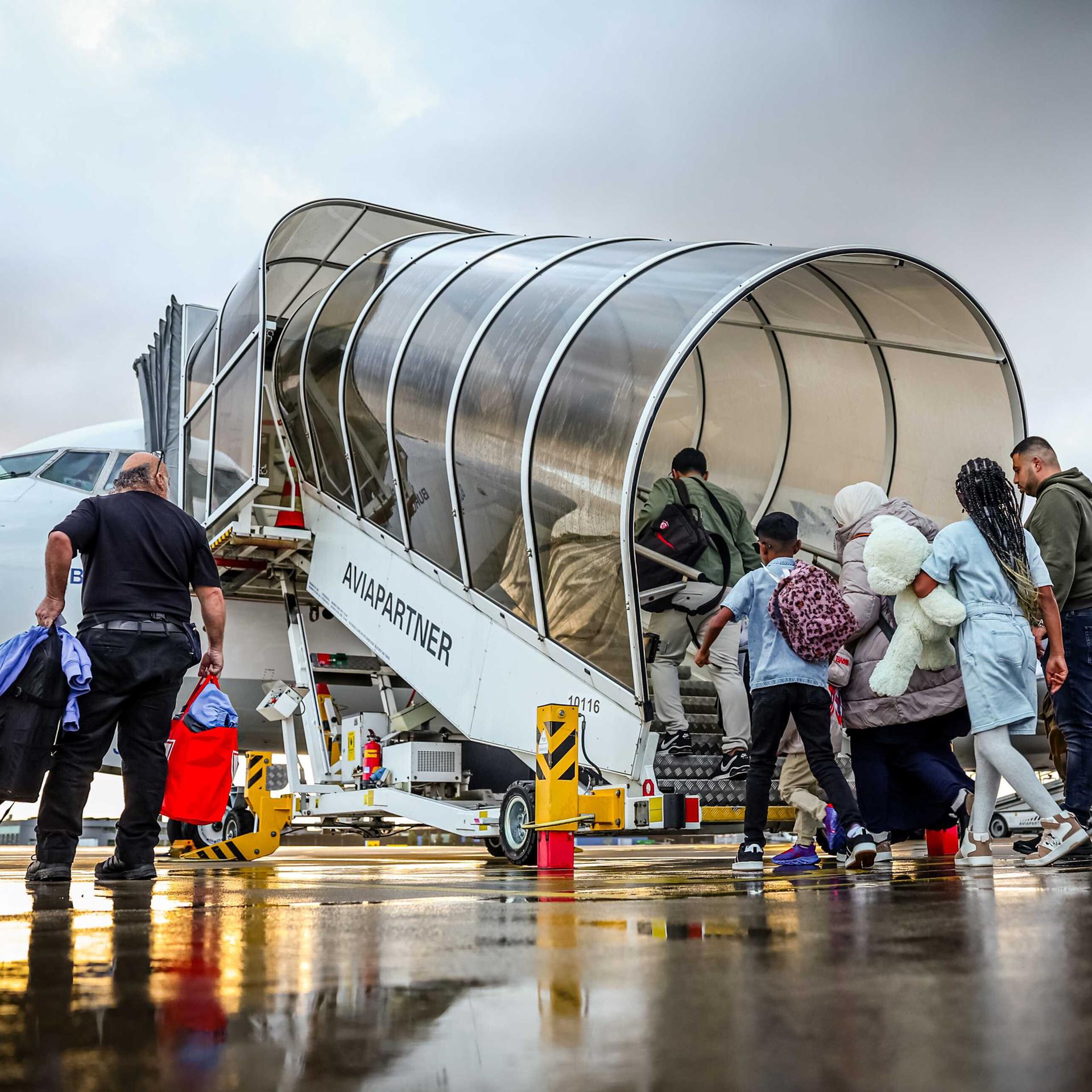 Blick auf eine Schlange von Menschen, die auf einem Rollfeld über eine überdachte Treppe in ein Flugzeug steigen. 