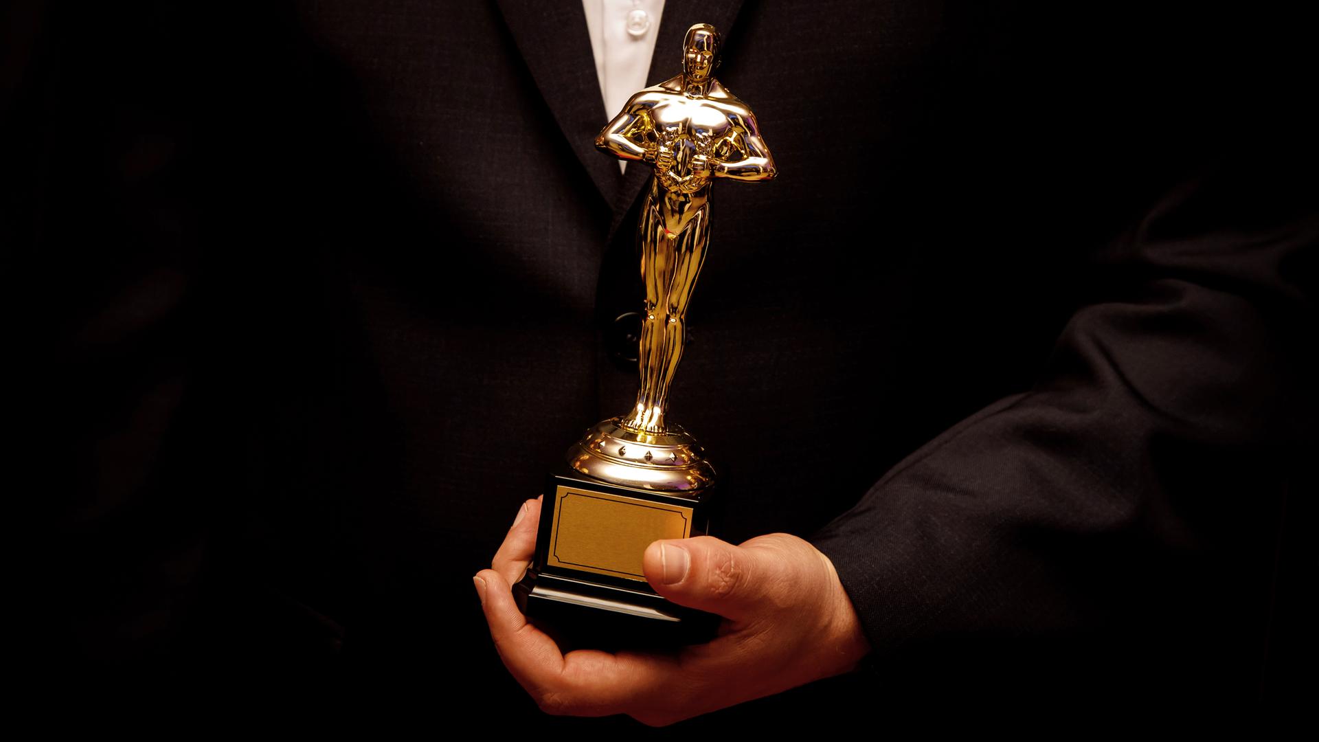Eine goldene Oscarstatue in der Hand von einem Menschen im schwarzen Anzug und weissem Hemd.
