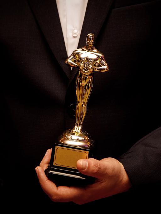 Eine goldene Oscarstatue in der Hand von einem Menschen im schwarzen Anzug und weissem Hemd.