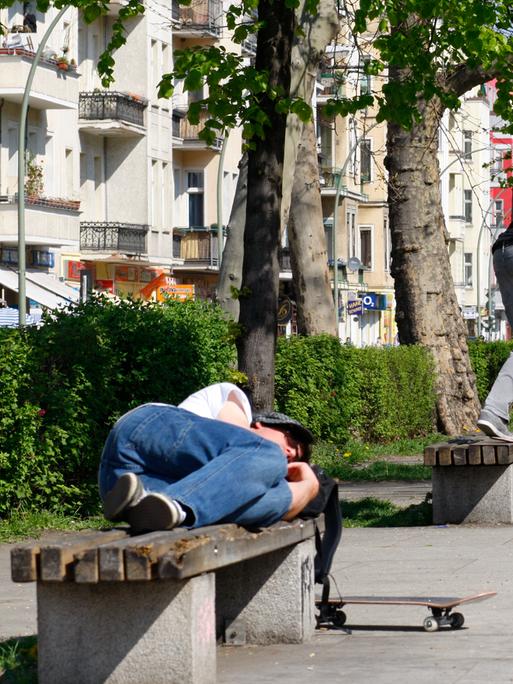 Ein Skateboarder schläft auf einer Parkbank während ein weiterer Skateboard hinter ihm auf einer Parkbank einen Trick aufführt.