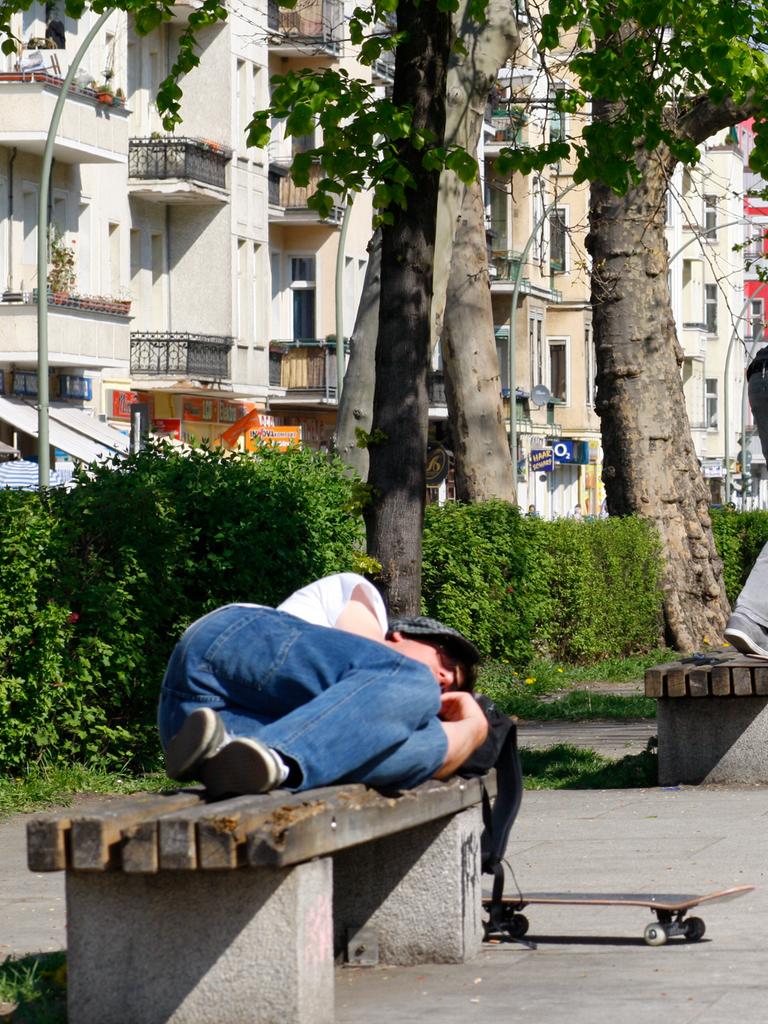 Ein Skateboarder schläft auf einer Parkbank während ein weiterer Skateboard hinter ihm auf einer Parkbank einen Trick aufführt.