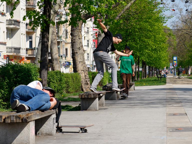 Ein Skateboarder schläft auf einer Parkbank während ein weiterer Skateboard hinter ihm auf einer Parkbank einen Trick aufführt.
