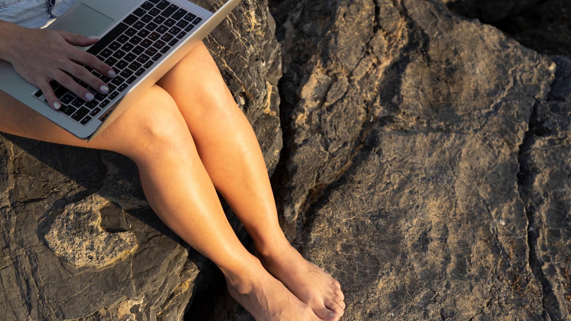 Eine Frau sitzt an einem Strand und hat einen Laptop auf den Knien.