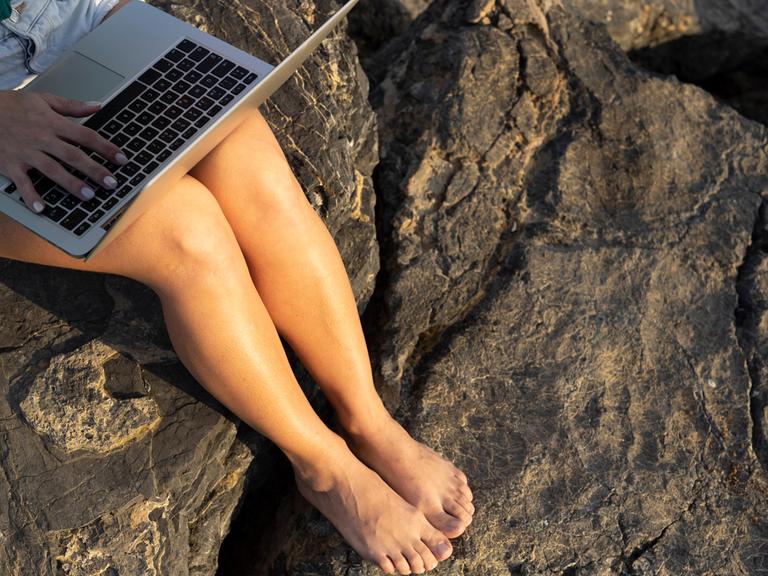 Eine Frau sitzt an einem Strand und hat einen Laptop auf den Knien.