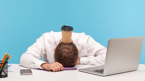Ein Mann hat das Gesicht auf den Schreibtisch gelegt, auf seinem Hinterkopf balanciert er einen Kaffee-to-go-Becher.
