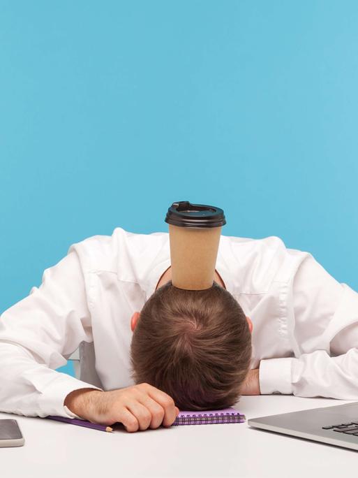 Ein Mann hat das Gesicht auf den Schreibtisch gelegt, auf seinem Hinterkopf balanciert er einen Kaffee-to-go-Becher.