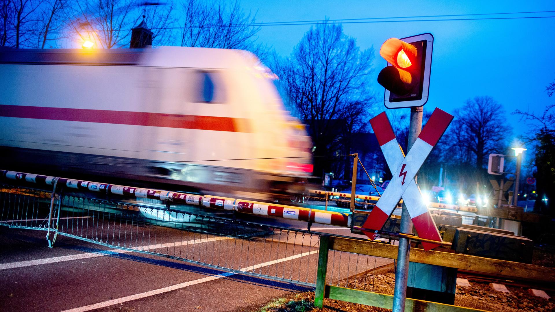Ein Andreaskreuz und eine rot leuchtende Ampel stehen an einem geschlossenen Bahnübergang während ein Zug der Deutschen Bahn passiert. 