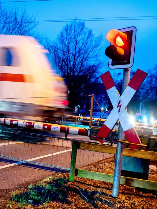 Ein Andreaskreuz und eine rot leuchtende Ampel stehen an einem geschlossenen Bahnübergang während ein Zug der Deutschen Bahn passiert. 