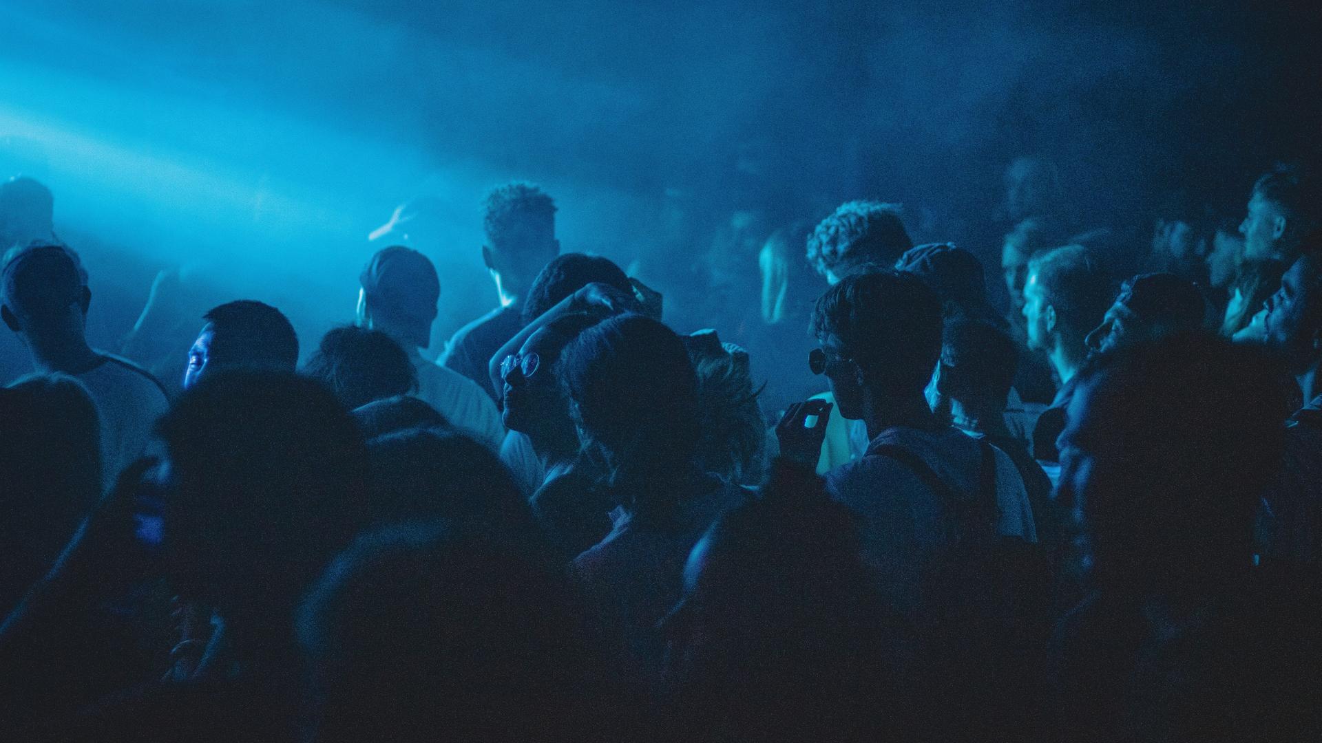 Club in blauem Licht in dem dicht gedrängt Menschen tanzen.