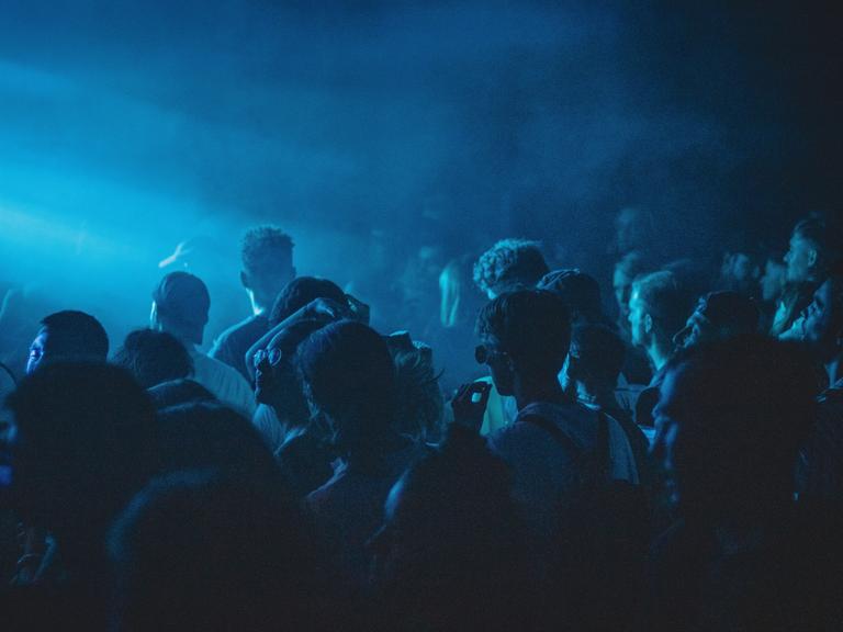 Club in blauem Licht in dem dicht gedrängt Menschen tanzen.